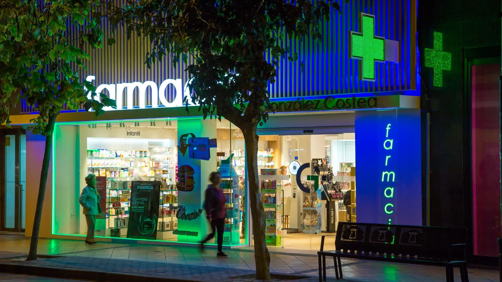 Exterior de una farmacia iluminada por la noche con cruces de farmacia verdes, letreros luminosos y una persona pasando por delante de la entrada.