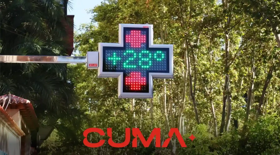 Logo de CUMA - Cruz de farmacia LED en una calle repleta de arboles con un follaje verde abundante.